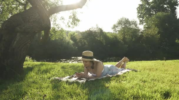 Женщина отдыхает в парке, слушает музыку из наушников и наслаждается летними каникулами. Милая очаровательная девушка в шляпе и платье лежит на одеяле на лугу у реки и расслабляет — стоковое видео