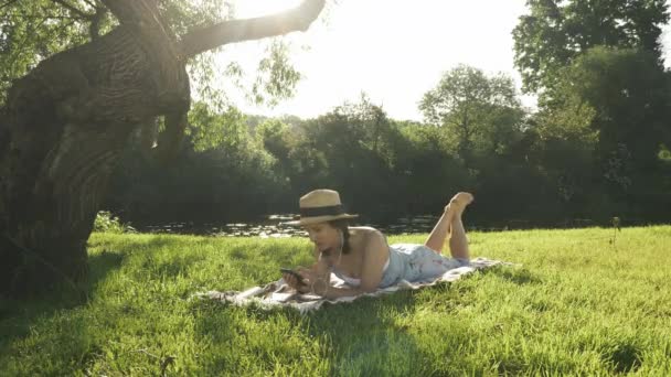Jolie jeune femme dans les écouteurs à l'écoute de la musique, profiter d'une chaude journée ensoleillée en plein air et se détendre dans le parc au bord de la rivière. Charmante femme couchée sur l'herbe sous un grand arbre et bavarder en ligne sur smartphone — Video