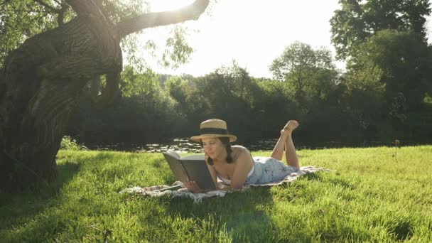 Encantadora joven con sombrero de paja y vestido acostado sobre una manta en la hierba en la naturaleza del parque, libro de apertura y empezando a leer. Morena linda mujer relajante al aire libre cerca del río — Vídeo de stock