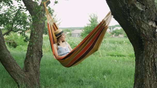Молода жінка лежить на гамаку серед двох дерев на відкритому повітрі. Милі дівчачі мрії, що лежать на гамаку в сільській місцевості. Брюнетка жінка в капелюсі і плаття розслабляється на гамаку зовні — стокове відео