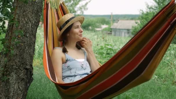 Heureuse femme joyeuse reposant dans l'hamac au jardin vert le week-end. Jeune femme couchée dans un hamac et profitant de vacances d'été à la campagne. Brunette femelle manger de la pomme et se détendre dans l'hamac — Video