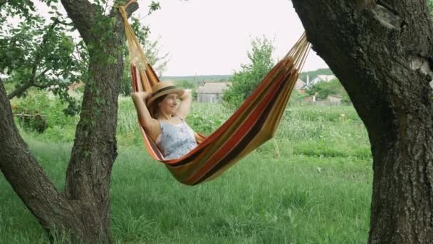 Jeune fille brune mignonne reposant sur un hamac, se détendre sur un hamac à la campagne. Femme caucasienne en chapeau et robe relaxant et levant les mains tout en étant couché sur hamac à l'extérieur — Video