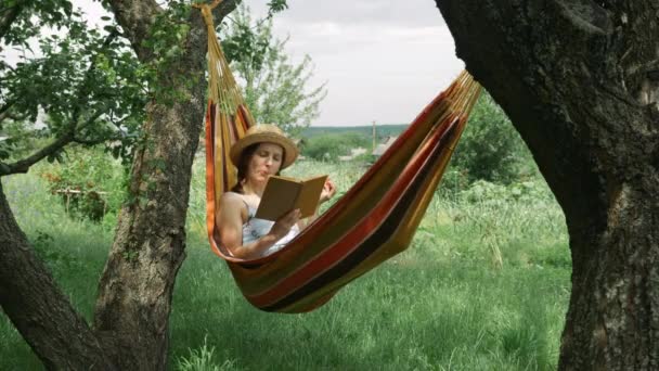 Ung brunett kvinna vilar i hängmatta mellan två träd på grön trädgård. Söt charmig flicka i hatt liggande i hängmatta, läsa bok och äta äpple utomhus. Sommarsemester på landet — Stockvideo