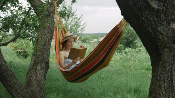 Donna che si rilassa in amaca nel giardino verde. Donna caucasica sdraiata sull'amaca, che legge libri e riposa in campagna. Ragazza in cappello e vestito in vacanza al villaggio. Donna con libro in amaca — Video Stock