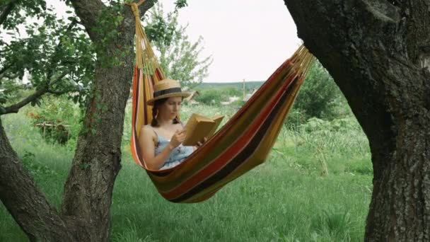 ผู้หญิงสาวที่มีหมวกพักผ่อนในเปลญวนที่สวนสีเขียว หนังสือการอ่านผู้หญิงสีน้ําตาลน่ารักที่มีเสน่ห์อยู่กลางแจ้งเปลญวน ผู้หญิงผ่อนคลายในเปลญวนกับหนังสือ — วีดีโอสต็อก