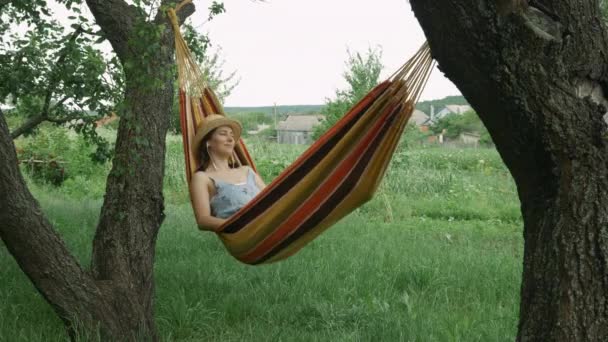 Jeune femme dans les écouteurs écouter de la musique dans un hamac, se reposer et se détendre. Femme brune en chapeau de paille écoute la musique des écouteurs tout en étant couché dans un hamac à l'extérieur — Video