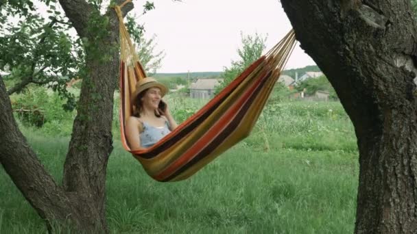 Femme brune parlant par téléphone portable dans un hamac à l'extérieur. Jeune fille parlant par smartphone tout en étant couché dans un hamac entre deux arbres dans un lieu de campagne confortable — Video