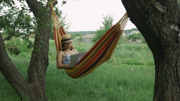 Jeune femme pigiste couché dans un hamac avec ordinateur portable à l'extérieur. Mignonne charmante femme en robe et chapeau assis dans un hamac à la nature et travaillant avec un ordinateur portable. Fille en utilisant un ordinateur portable couché dans un hamac — Video