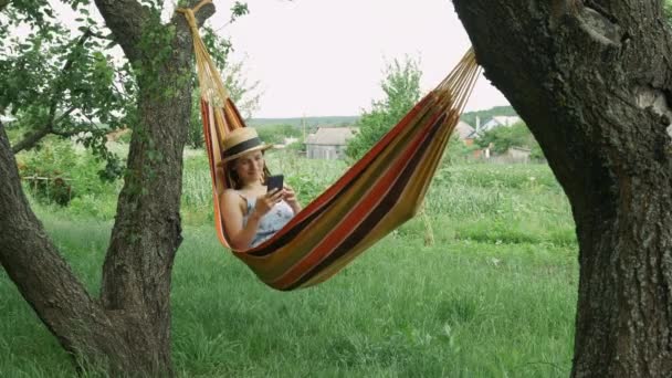 Jeune femme détendue regardant le téléphone mobile dans l'hamac dehors. Charmante brune couchée dans un hamac entre deux arbres à la campagne. Fille en chapeau et robe en utilisant le smartphone dans l'hamac — Video