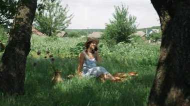 Kırsal kesimde çimenlerde oturan ve akıllı telefondan konuşan bir kadın. Şapkalı ve elbiseli genç esmer kadın yeşil bahçede cep telefonuyla konuşuyor.