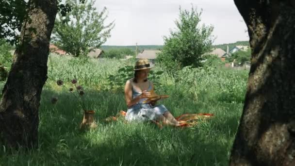 Mulher caucasiana atraente sentado na grama no jardim verde e livro de leitura no verão dia ensolarado. Menina encantadora bonito em chapéu e vestido com livro em mãos no parque. Feminino livro de leitura ao ar livre — Vídeo de Stock