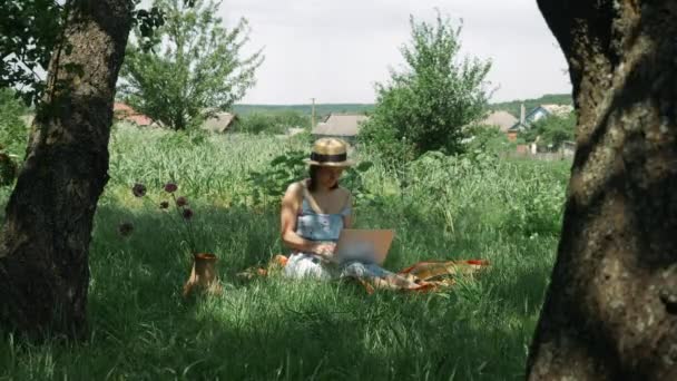 Green Garden 'da dizüstü bilgisayarda çalışan serbest çalışan bir kadın. Şapkalı ve elbiseli genç esmer kadın parkta çimlerde oturuyor ve dizüstü bilgisayar kullanıyor. Defteri açık havada olan kız. — Stok video