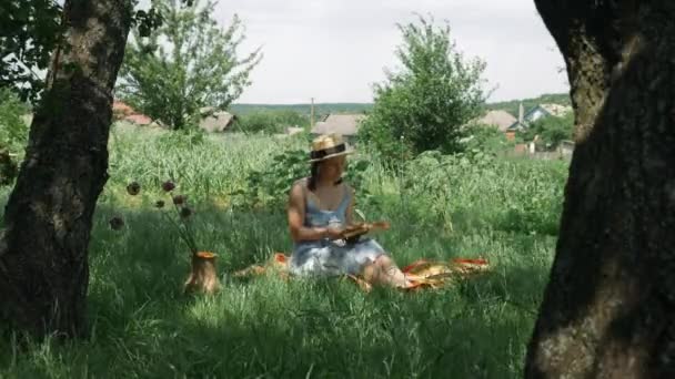 Weibliches Lesebuch im grünen Garten. Junge charmante Frau mit Hut und Kleid sitzt auf dem Gras im Park und liest Buch, entspannt und genießt den Sommerurlaub. Mädchen mit Buch im Freien — Stockvideo