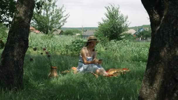 Frau mit Kopfhörer sitzt auf Gras im grünen Garten und hört Musik. Glücklich lächelnde Frau mit Hut und Kleid hört Musik aus Kopfhörern, singt und entspannt im grünen Park — Stockvideo