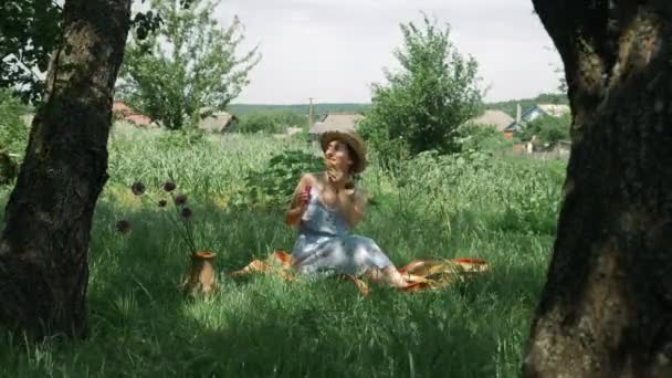 Женщина пускает пузыри в зеленый сад. Женщина на пикнике. Молодая милая девушка в платье и шляпе сидит на клетку в зеленом парке и дует пузырьки. Брюнетка отдыхает в парке — стоковое видео