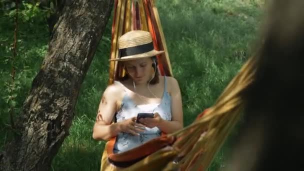 Vrouw in oortjes liggend in hangmat en luisterend naar muziek in de groene tuin. Brunette vrouw luistert naar muziek van oortelefoons, met behulp van mobiele telefoon en het zingen van het lied in hangmat. Meisje ontspannen in hangmat — Stockvideo