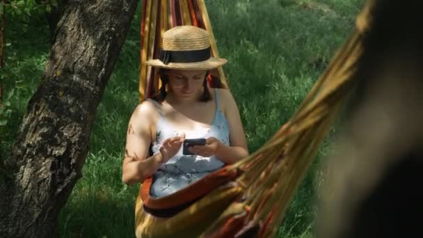 Femme avec téléphone portable couché dans un hamac. Jeune femme reposant dans un hamac avec smartphone dans les mains. Brunette fille en chapeau en utilisant un téléphone portable tout en se relaxant dans l'hamac à l'extérieur — Video