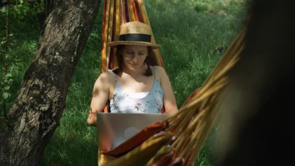 Jeune femme brune dans un chapeau couché dans un hamac et travaillant sur un ordinateur portable au jardin vert. Charmante femme reposant dans l'hamac et utilisant l'ordinateur portable à l'extérieur. Concept vacances d'été — Video