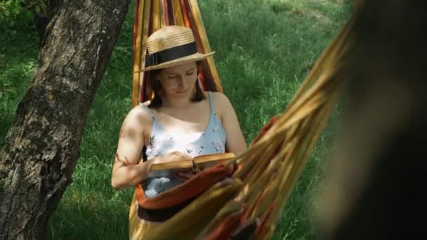 ハンモックで本を持つ帽子の若い女性。ハンモックで休んで緑の庭で本を読んでいる魅力的な幸せな女性。ブルネット女性嘘でハンモックで手に本、リラックスして休暇を楽しむ — ストック動画