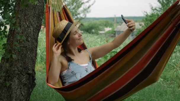 Joven mujer feliz tomando foto selfie con el teléfono móvil acostado en la hamaca. Encantadora morena femenina relajándose en la hamaca y toma fotos selfies en el teléfono inteligente — Vídeo de stock