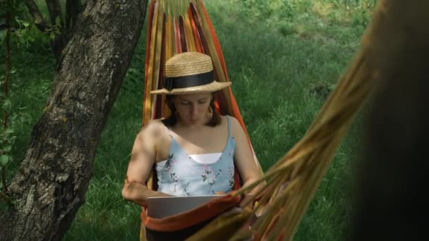 Femme dans le chapeau reposant sur l'hamac et travaillant sur l'ordinateur portable au jardin vert. Femme utilisant l'ordinateur portable dans l'hamac dehors. Femme pigiste travaillant sur ordinateur portable portable portable couché dans un hamac à la campagne — Video