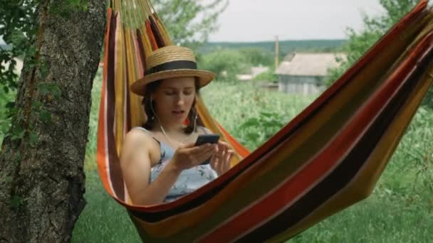 Femme dans des écouteurs couchés dans un hamac et écoutant de la musique, chantant et se relaxant dans un jardin verdoyant. Jolie fille souriante dans le chapeau et la robe reposant dans l'hamac et écoute la musique des écouteurs — Video