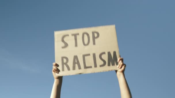 Plakat STOP RACISM vor blauem Himmel. Hände gegen den blauen Himmel mit dem Schild "Stop Racism". Kundgebungen gegen Polizeibrutalität und Pluralität der Gewalt. Friedlicher Protest unter dem Motto "Black Lives Matter" — Stockvideo