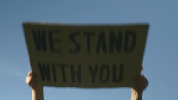 女性の手は青い空に対してあなたと一緒に立つサインを上げる。女性はポスター"We Stand With You"を持っている。黒人の生活は平和的な抗議である。警察の残虐行為や反黒人人種差別に反対する集会 — ストック動画