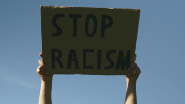 Женщина держит плакат "Остановите расизм". Подпишите "Остановить расизм" против голубого неба. Плакат "Остановите расизм" в руках кавказской женщины. Митинги против жестокости полиции и множественности насилия — стоковое видео