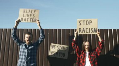 Siyah Yaşamlar Madde 'yi ve mavi gökyüzüne karşı RACISM' i durduran posterlerle ilahi söyleyen insanlar. Siyahi karşıtı ırkçılığa ve şiddetin çoğulculuğuna karşı barışçıl protestolarda kadın ve erkek tabelaları var.