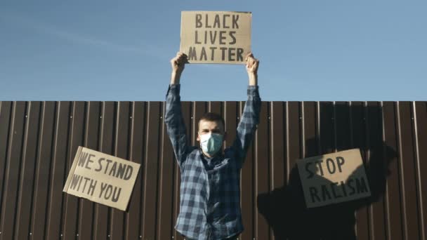 Homem caucasiano em máscara protetora cantando com sinal "Vidas negras importam" contra a parede marrom e céu azul. Homem acenando BLACK LIVES MATTER cartaz de protesto fora. Parem com o racismo e a brutalidade policial — Vídeo de Stock