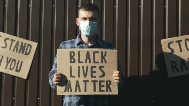 戴防护面罩的男人拿着海报"黑人的生命很重要"，背景是棕色的。在反对警察暴行的集会上，拿着标有"黑人生命很重要"的白种人年轻人手拉手。制止种族主义 — 图库视频影像