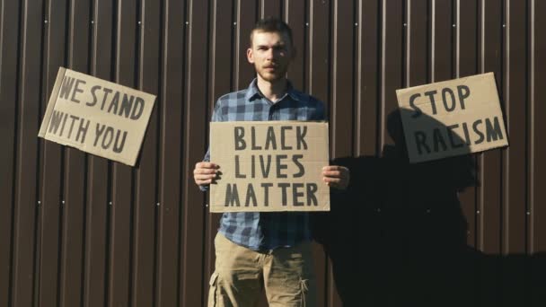 Millennial man met bord "zwarte levens materie" tegen bruine muur. Stop Racisme. Poster BLACK LIVES MatTER in handen van een blanke man bij een demonstratie tegen politiegeweld en pluraliteit van geweld — Stockvideo