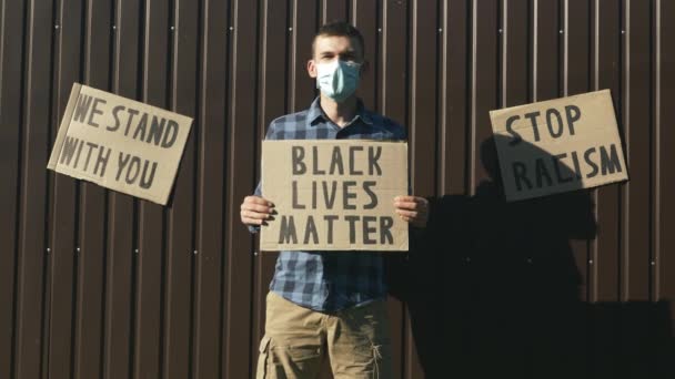 Homem em máscara protetora com sinal "Vidas negras importam" contra a parede marrom com sinais "Estamos com você", "Sem racismo". Comícios contra a brutalidade policial. Protesto pacífico — Vídeo de Stock