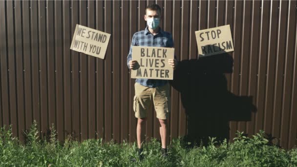 Giovane uomo millenario in maschera protettiva in piedi contro sfondo marrone con poster "Black vive la materia" nelle mani. L'uomo caucasico tiene il segno "Le vite nere contano". Rallies contro la brutalità della polizia — Video Stock