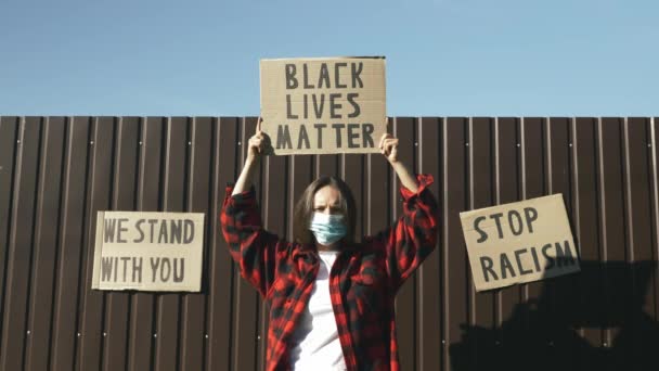 Kavkazská žena drží plakát Černá hmota žije nad hlavou na hnědém pozadí s nápisy Stojíme při vás, Žádný rasismus. Žena v ochranné masce s nápisem BLACK LIVES MATTER v rukou