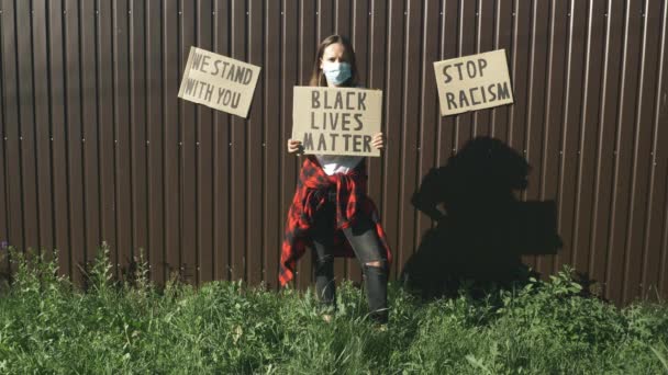Młoda tysiącletnia dziewczyna w ochronnej masce twarzy stojąca przed brązową ścianą z plakatem BLACK LIVES ma znaczenie na wiecu o wielości przemocy i anty-czarnym rasizmie. Pokojowy protest Black Lives Matter — Wideo stockowe