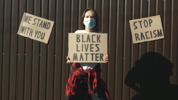 Wanita kaukasia dengan topeng wajah pelindung dengan poster "Kehidupan kulit hitam penting" di tangan dengan latar belakang coklat. Perempuan berdiri menghadap dinding coklat dengan tanda "Kehidupan Hitam itu penting". Hentikan konsep rasisme — Stok Video