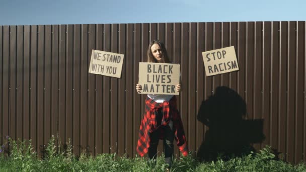 Junge kaukasische Millennial-Frau mit Schild "Black lives matter" an Wand mit Schildern "Kein Rassismus", "Wir stehen an eurer Seite". Stop Racism Konzept. Schild "BLACK LIVES MATTER" in den Händen einer kaukasischen Frau — Stockvideo