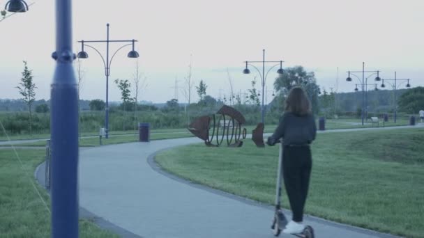 幸せな千年紀の少女は日没時に都市の遊歩道で生態電動スクーターに乗っている。白人女性は仕事から家庭への個人的なエコ輸送に乗る。都市の健康と生態系のライフスタイル — ストック動画