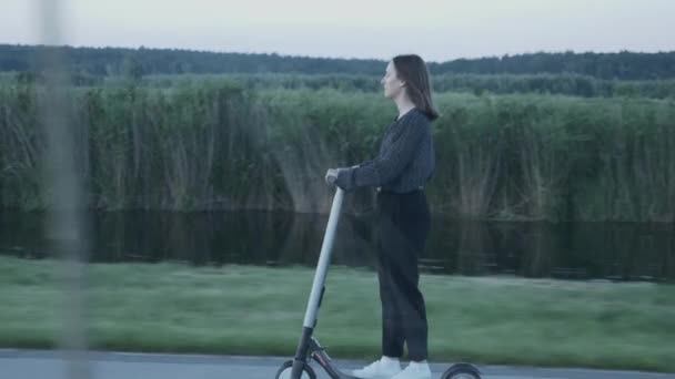 A seguire scatto laterale di giovane donna moderna alla guida di scooter elettrico personale al tramonto. Ragazza cavalca scooter elettrico moderno lungo la strada in parco. Trasporti ecologici e moderni nelle città — Video Stock