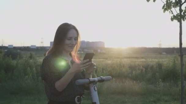 Retrato de una exitosa empresaria con teléfono móvil y scooters eléctricos en el paseo marítimo de la ciudad al atardecer. Mujer con smartphone en las manos de pie cerca de e-scooter cargado en la calle. Transporte eléctrico — Vídeos de Stock