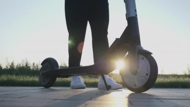 Donna che inizia a cavalcare su scooter elettrico in città al tramonto. Primo piano della donna che guida e-scooter in estate. Ragazza cavalca carico scooter elettrico lungo la strada in parco al tramonto. Trasporto elettrico — Video Stock