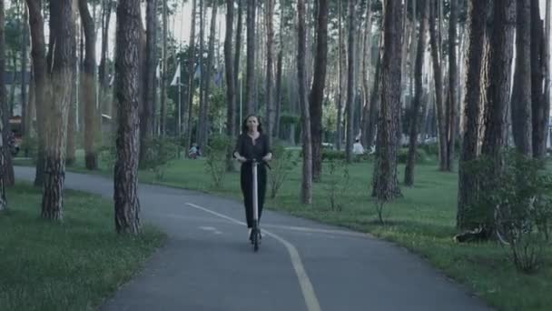 Fille promenades scooter électrique ou e-scooter dans le parc de la ville au coucher du soleil. "Ecological e-mobility in urban city". Femme en costume équitation électrique kick scooter dans le parc. Femme utilisant le transport électrique dans un parc urbain — Video