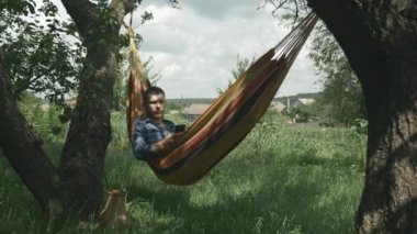 Bahçedeki hamakta cep telefonuna bakan rahat genç adam. Erkek, açık havada hamakta yatıyor. Hamakta akıllı telefon kullanan adam