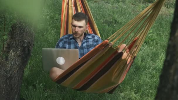 Um homem a trabalhar num portátil deitado numa rede ao ar livre. Freelancer masculino trabalha no computador portátil em férias de verão no campo. Homem relaxante em rede — Vídeo de Stock