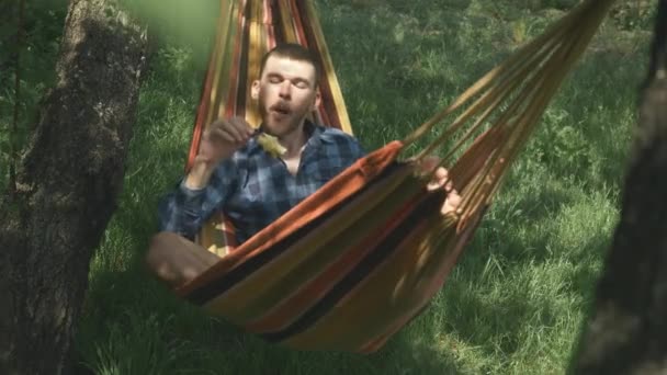 ハンモックでリラックス。ハンモックに寝そべってリンゴを食べる若い屈託のない男。田舎のハンモックでリラックスした男性観光客。休暇を屋外で楽しんでいる男。観光キャンプ。夏休み — ストック動画