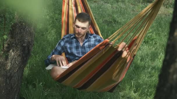 Молодой человек открывает ноутбук и начинает работать, лежа в гамаке на открытом воздухе. Человек, работающий над записной книгой в зеленом саду. Свободное рабочее место. Летний туристический лагерь — стоковое видео