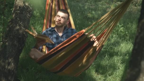 Campeggio estivo turistico. Turista maschile che si rilassa in amaca in vacanza. Uomo sdraiato sull'amaca e a leggere libri. Uomo godendo fine settimana all'aperto nella giornata di sole. Rilassarsi in amaca — Video Stock