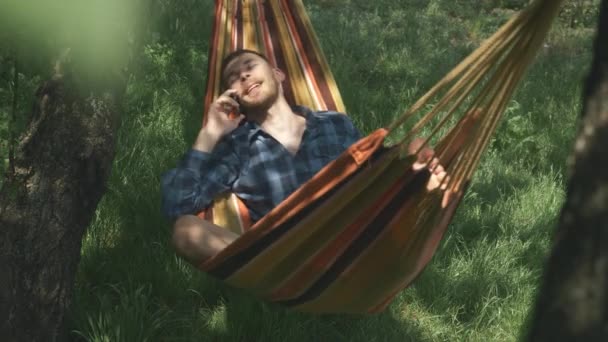 ハンモックに横たわっている携帯電話で話すハンサムな幸せな男。男性は携帯電話で話し、ハンモックで揺れる。ハンモックでリラックス。夏休みだ。キャンプ — ストック動画